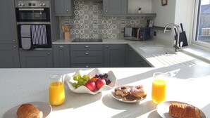 A modern kitchen with grey cabinets, a white countertop, and a breakfast setting with croissants, fruit, and orange juice. - Stylish Skipton Retreat | Private Garden | Parking | Hiking | Yorkshire Dales (Skipton)
