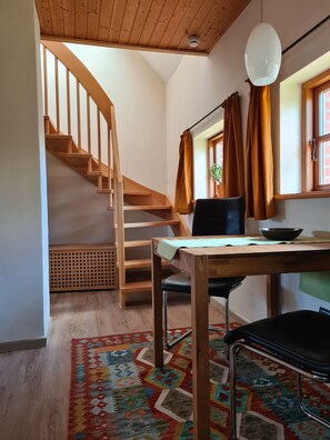 A room with a wooden staircase, a table with a chair, a hanging light fixture, and a window with curtains. - Vacation home **** star certified, 3 minutes walk from the Hasbruch (Hude (Oldb))