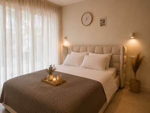 Une chambre à coucher avec un grand lit, une table de chevet avec une lampe, une horloge au mur et une fenêtre avec des rideaux légers.