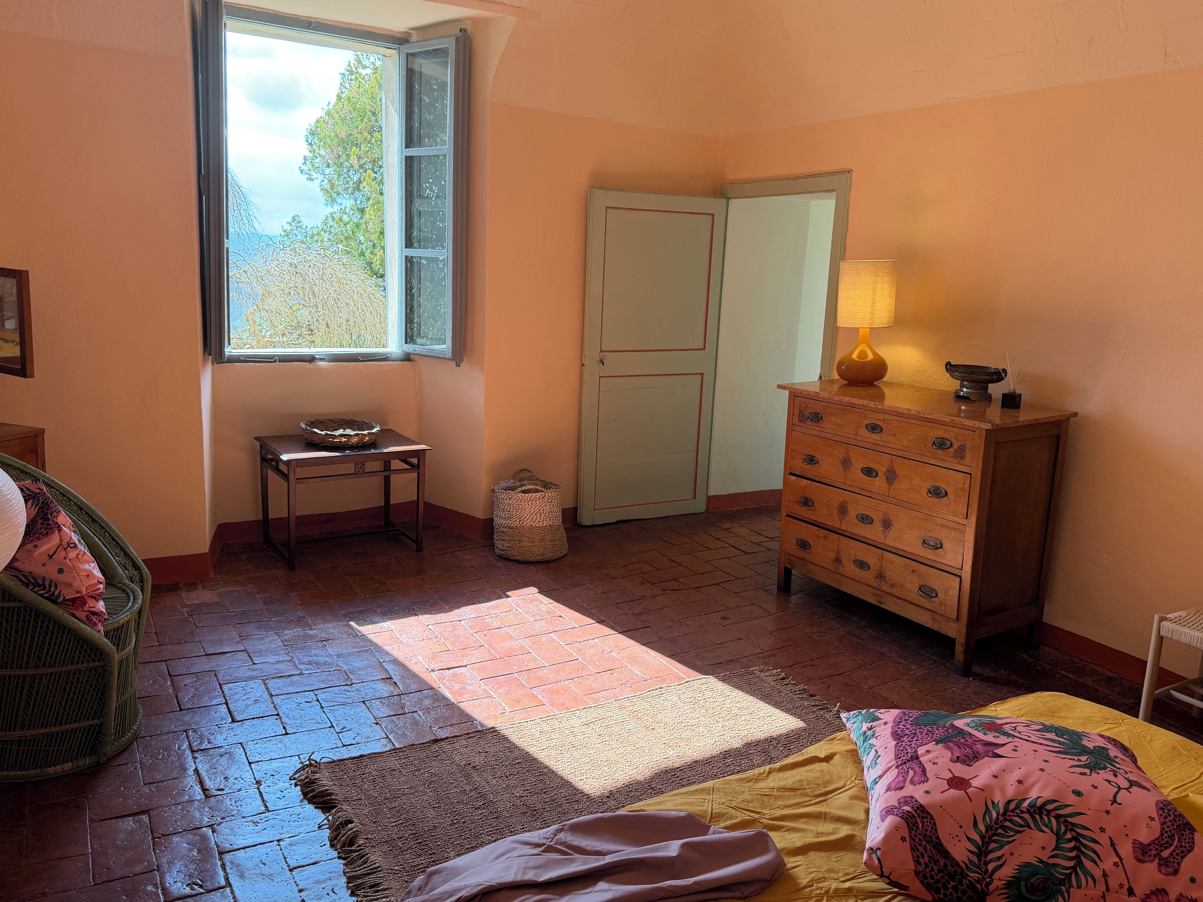 A room with a window, a wooden dresser, a wicker chair, and a bed with a patterned bedspread.