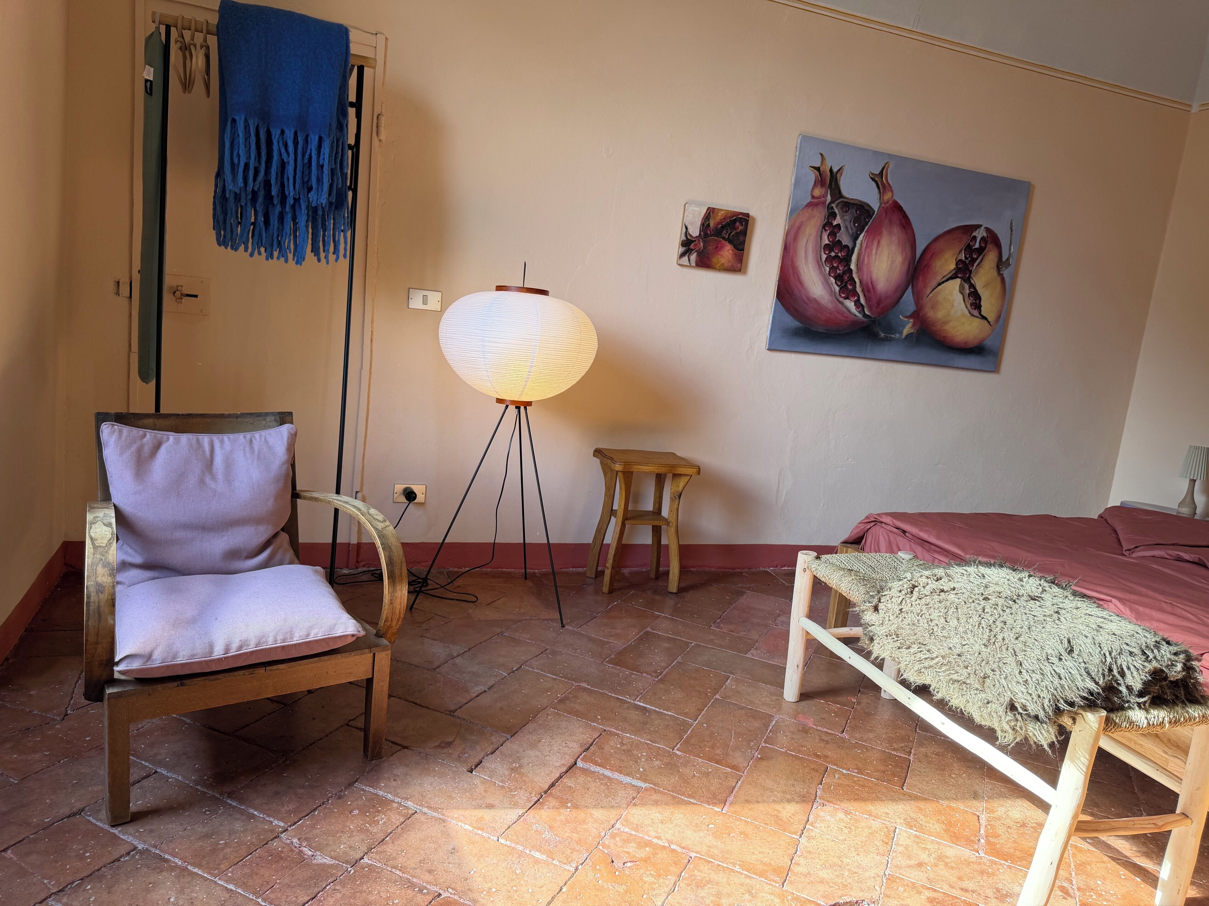 Une pièce comprenant une chaise, un lampadaire, un petit tabouret, un lit et un tableau représentant des grenades, accroché au mur.