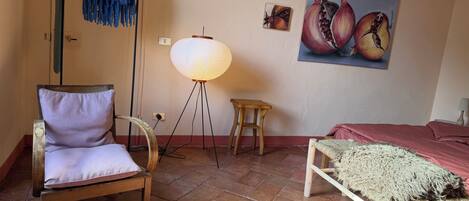 Une pièce comprenant une chaise, un lampadaire, un petit tabouret, un lit et un tableau représentant des grenades, accroché au mur.