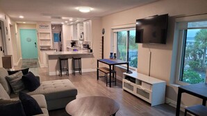 A modern living room with a sofa, a coffee table, a TV, and a kitchen area in the background. - Bahia Del Sol Condo (Ruskin)
