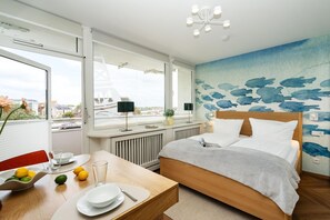 Une chambre d’hôtel avec un lit, une table à manger avec des assiettes et des verres, une fenêtre donnant sur des immeubles et un mur orné d’une fresque représentant une baleine bleue.