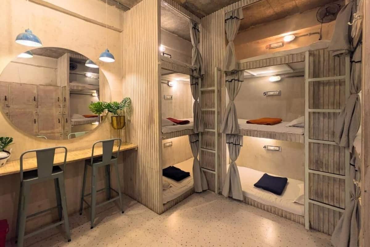 Quarto compacto com beliches, uma pequena escrivaninha com banquetas, um espelho e um vaso de planta.