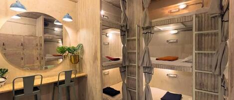 A compact room with bunk beds, a small desk with stools, a mirror, and a potted plant.