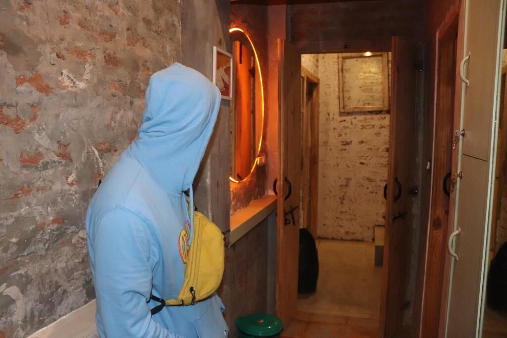 A person in a blue hoodie and yellow bag standing in a hallway with a rustic brick wall, a wooden door, and a neon light.