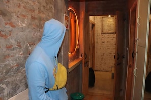 A person in a blue hoodie and yellow bag standing in a hallway with a rustic brick wall, a wooden door, and a neon light. - Bunkyard Hostel (Udaipur)