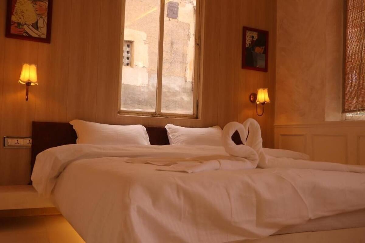 Quarto de hotel com uma cama, duas luminárias fixadas na parede, uma janela com vista para edifícios e um quadro na parede.