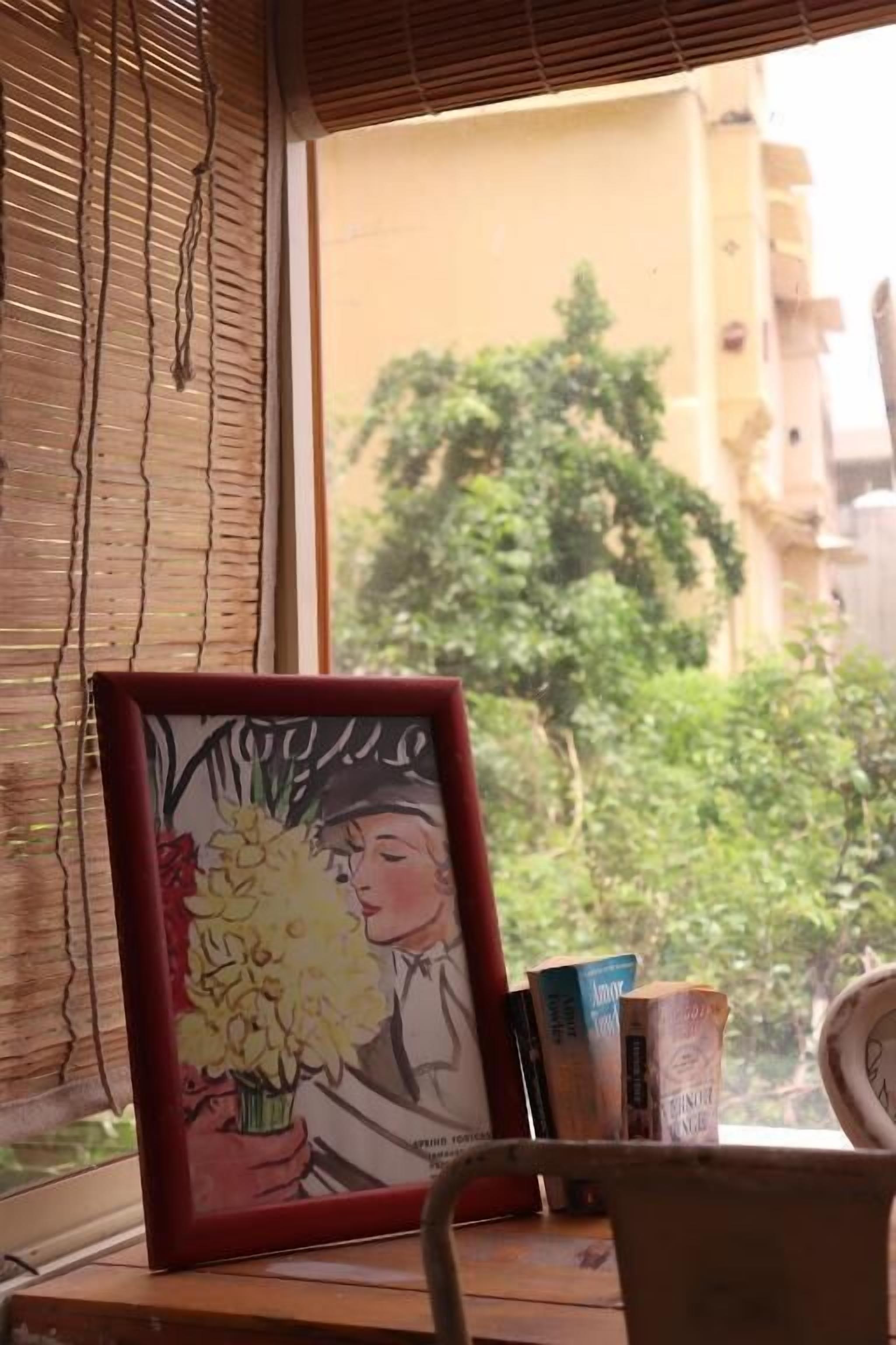 Una obra de arte enmarco que muestra a una persona sosteniendo flores sobre una mesa cerca de una ventana con persianas, libros y una silla.
