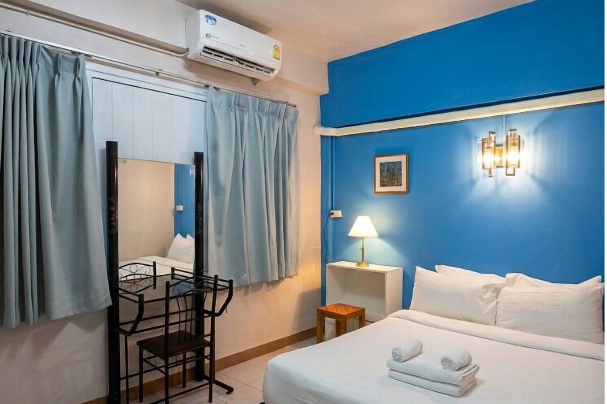 A hotel room with a bed, a desk with a chair, a small table with a lamp, a wall-mounted air conditioner, and a picture on the wall.