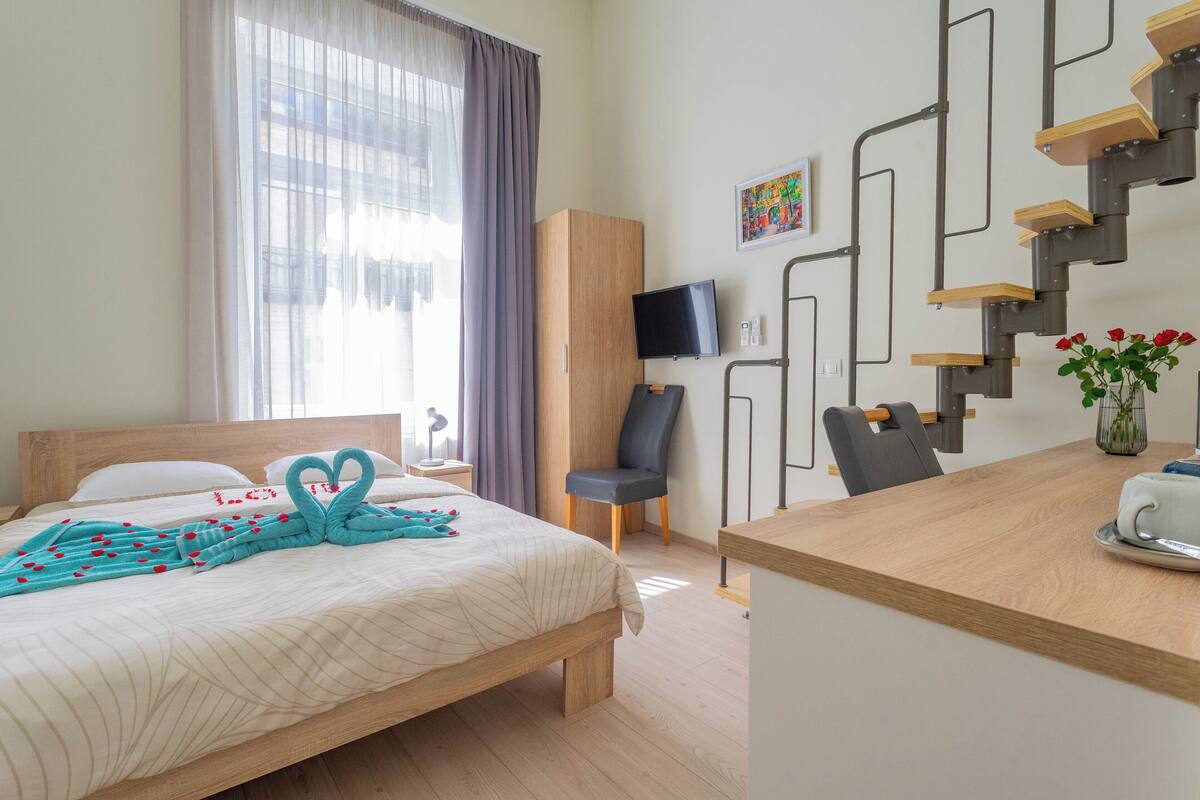 A hotel room with a bed, a desk, a chair, a TV, a staircase, and a window with curtains.
