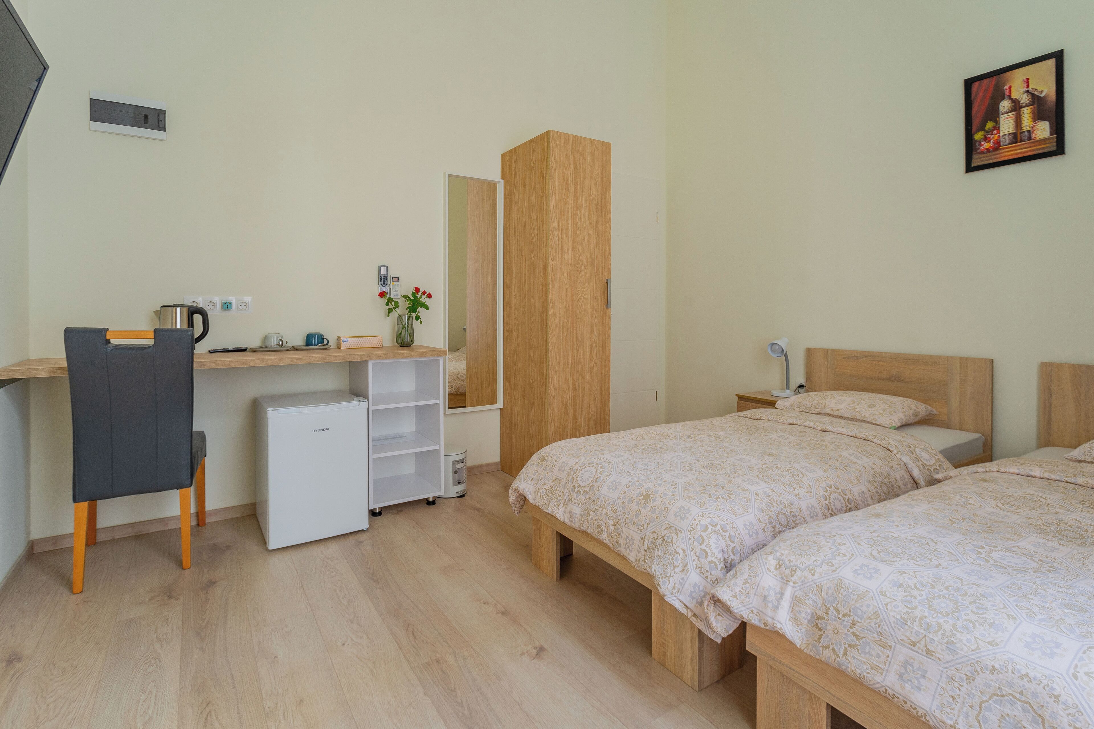 A bedroom with two beds, a desk with a chair, a wardrobe, and a small refrigerator.