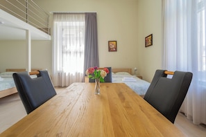 A hotel room with a wooden dining table, two black chairs, a bed with a white headboard, a window with sheer curtains, and a small vase with flowers on the table.