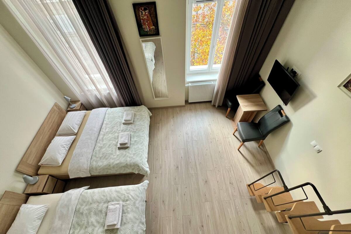 A hotel room with two beds, a desk, a chair, a TV, and a window with autumn leaves.