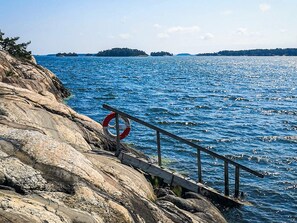 A rocky shoreline with a lifebuoy attached to a railing, overlooking a calm sea with distant islands. - 5 Star Holiday Home in Navekvarn (Navekvarn)