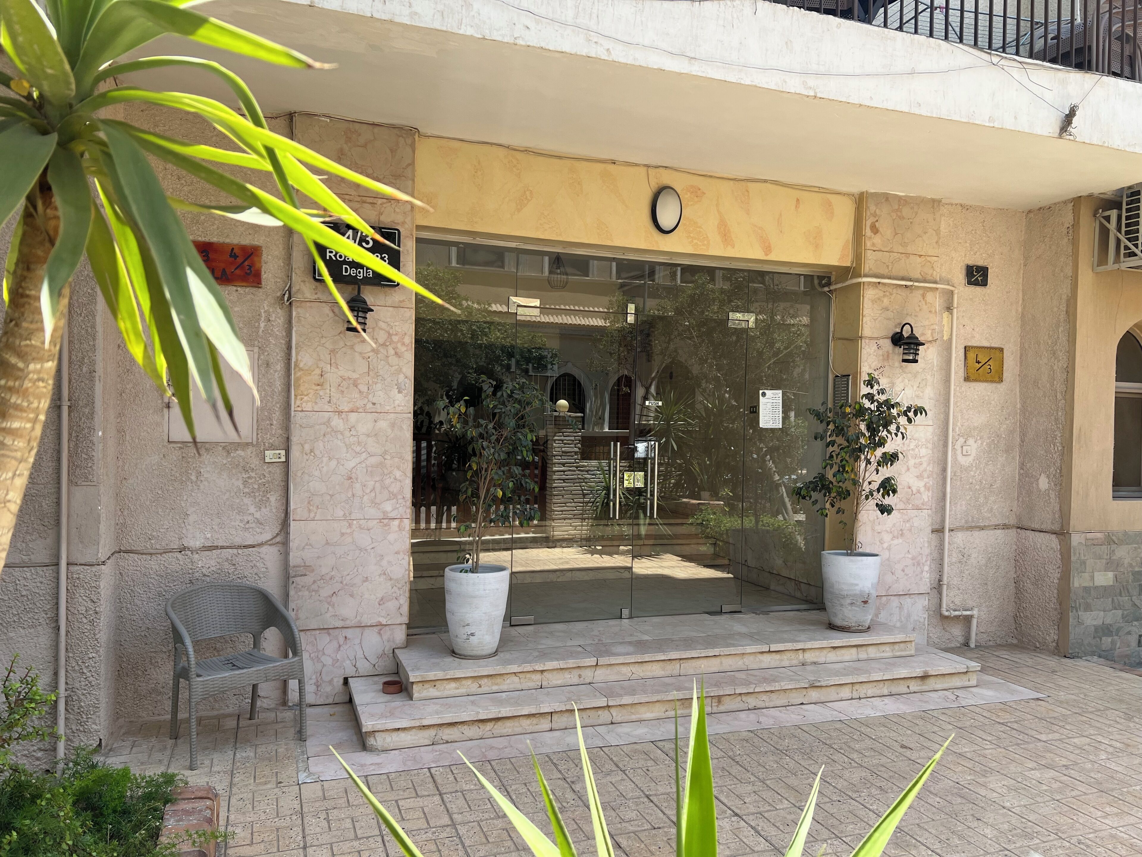 Une entrée d’immeuble avec une façade en pierre, un grand pot de fleurs, un banc et une porte en verre transparent.