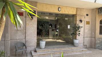 Une entrée d’immeuble avec une façade en pierre, un grand pot de fleurs, un banc et une porte en verre transparent.