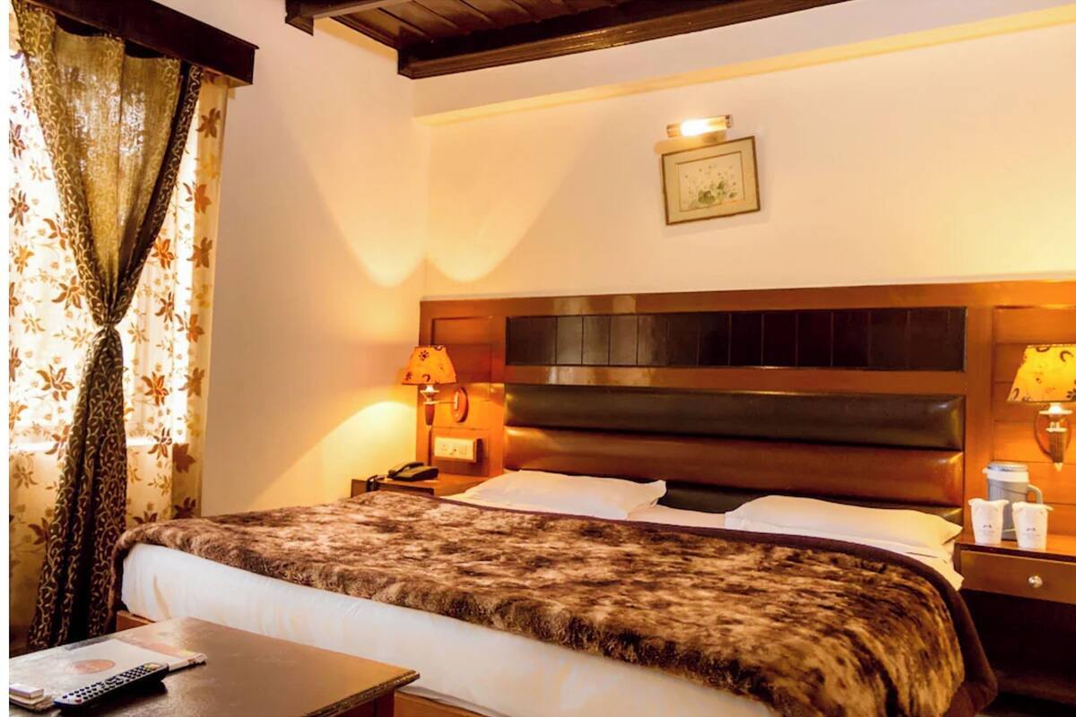 Habitación de hotel con una cama grande, dos lámparas de noche, una mesita de noche, un cuadro enmarcado en la pared y una ventana con cortinas.