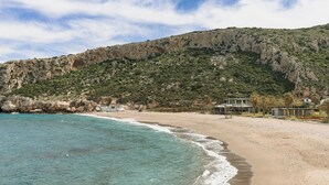 Een strand met helder turquoise water, een rotsachtige heuvel en enkele gebouwen in een eenvoudige bouwstijl.