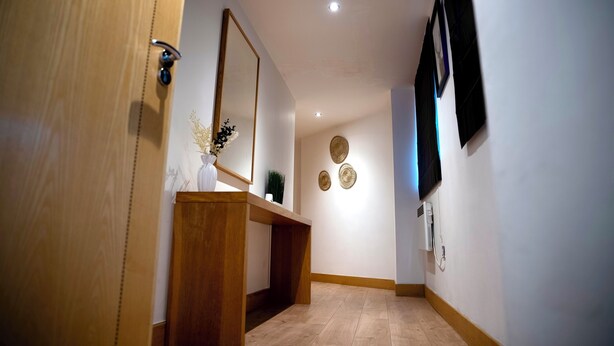 A hallway with a wooden door, a wooden console table with a vase, a mirror, a wall-mounted TV, and a wall with three decorative plates. - The Comfort Hub, Leeds Train Station (Leeds)