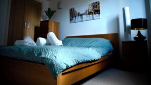 A bedroom with a wooden bed, a nightstand with a lamp, a wardrobe, and a painting on the wall. - The Comfort Hub, Leeds Train Station (Leeds)