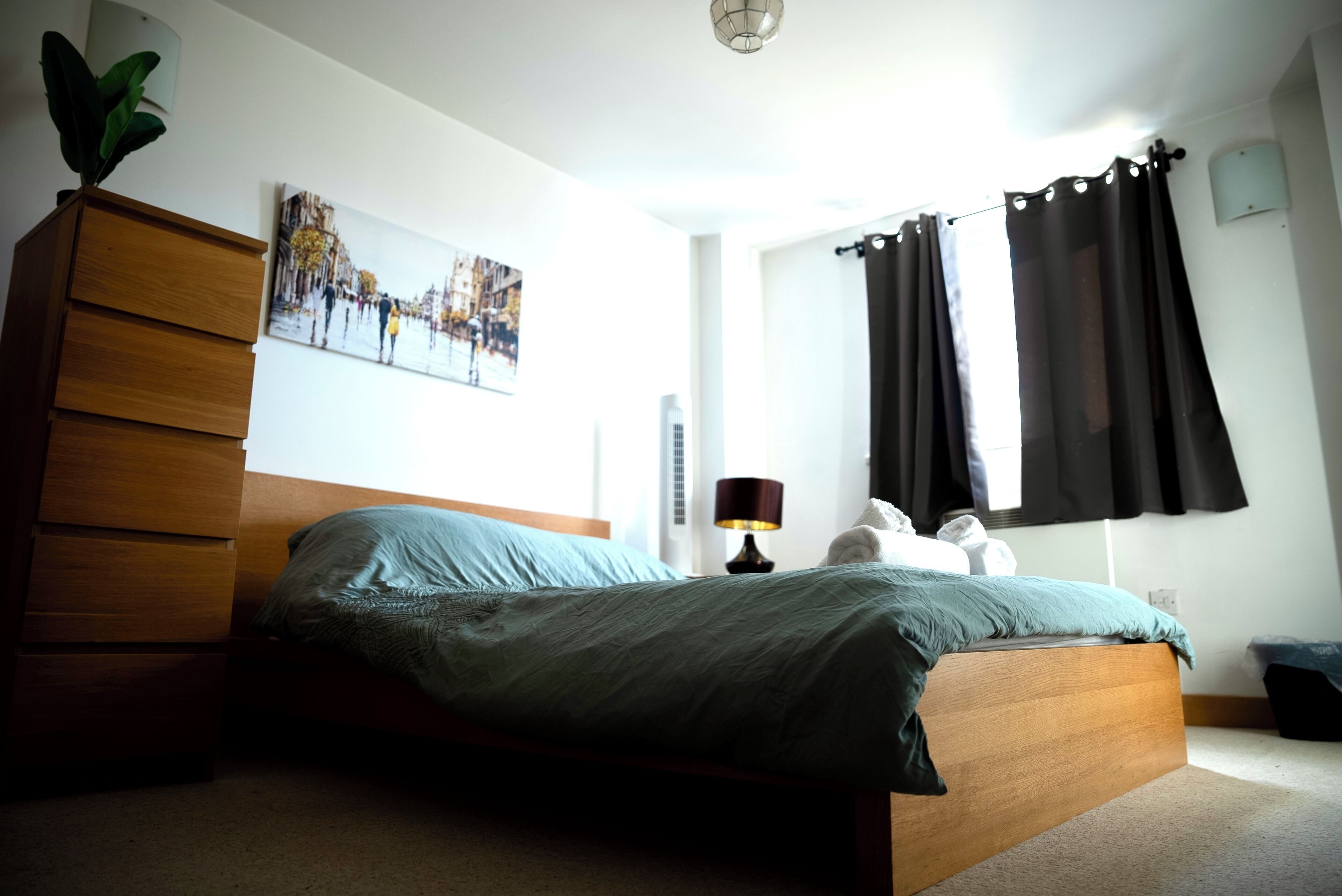 A bedroom with a bed, wooden dresser, lamp, and a cityscape painting on the wall.
