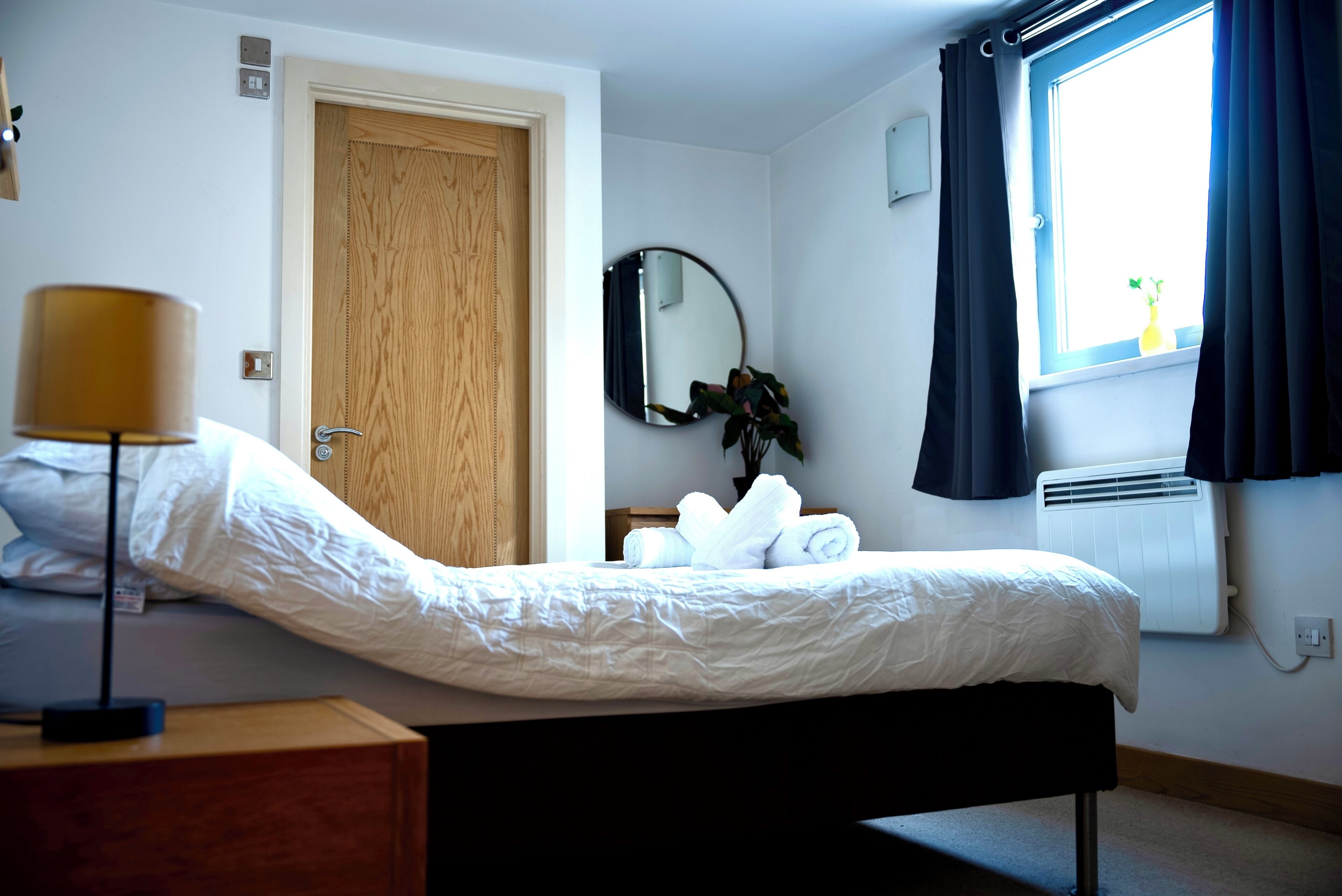 A hotel room with a bed, a bedside lamp, a mirror, a window with curtains, a radiator, and a plant.
