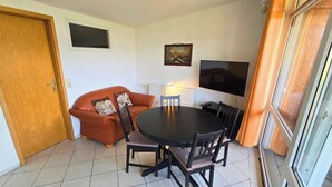 Uma sala de jantar com uma mesa redonda e cadeiras, um sofá, uma televisão e uma porta.