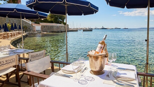 Une table dressée pour le dîner, avec une bouteille de champagne, de l’eau claire, et une vue sur des bateaux et un mur de pierre.