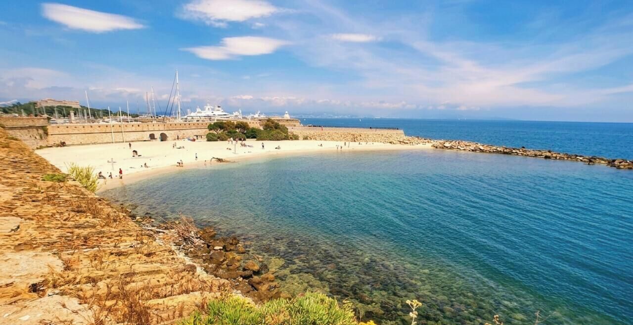 A coastal beach with clear water, a rocky shoreline, and a marina with boats.