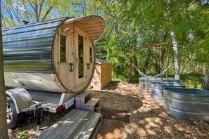 A wooden cabin with a large window, a metal trailer, and metal barrels in a wooded area.