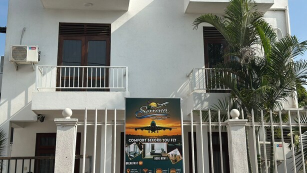 A two-story building with a white facade, a balcony with a railing, and a sign advertising an airport guest house. - Serrena Airport Guest House - Colombo (Seeduwa)