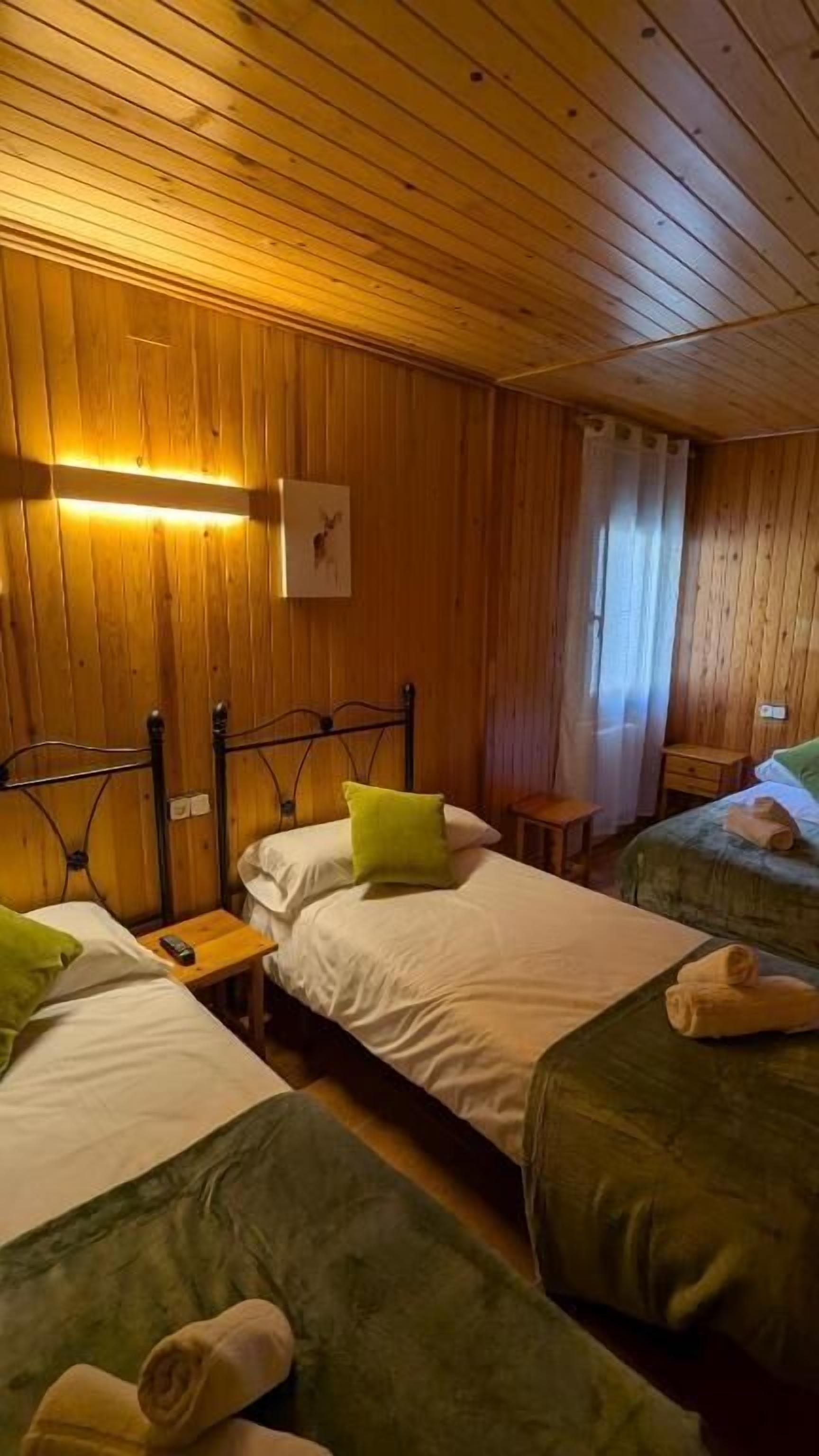 Habitación con paredes y techo de madera, dos camas con ropa de cama verde, una mesita pequeña y una mesita de noche.
