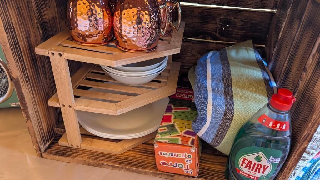 Une étagère en bois avec des tasses en céramique, un égouttoir, un coussin à rayures, une bouteille de détergent et une boîte colorée.