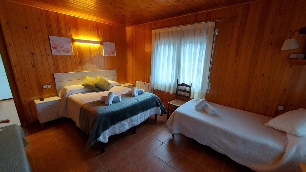 Una habitación con paneles de madera, dos camas, una silla, una mesita de noche y una ventana con cortinas.