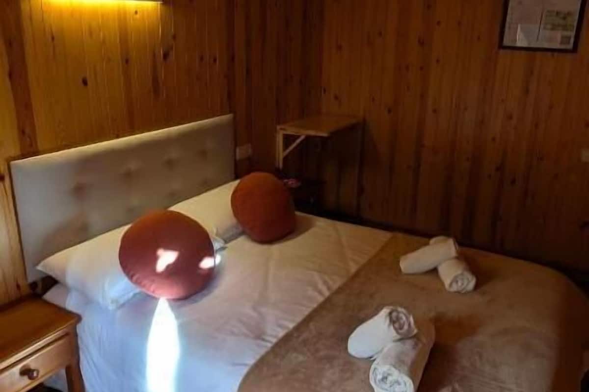 Une pièce aux murs lambrés, comprenant un lit, des oreillers, une table de chevet et une lampe fixée au mur.