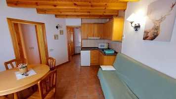 Una habitación acogedora con techo de madera, una mesa de comedor con sillas, un sofá, una zona de cocina con armarios, y una lámpara fijada a la pared.
