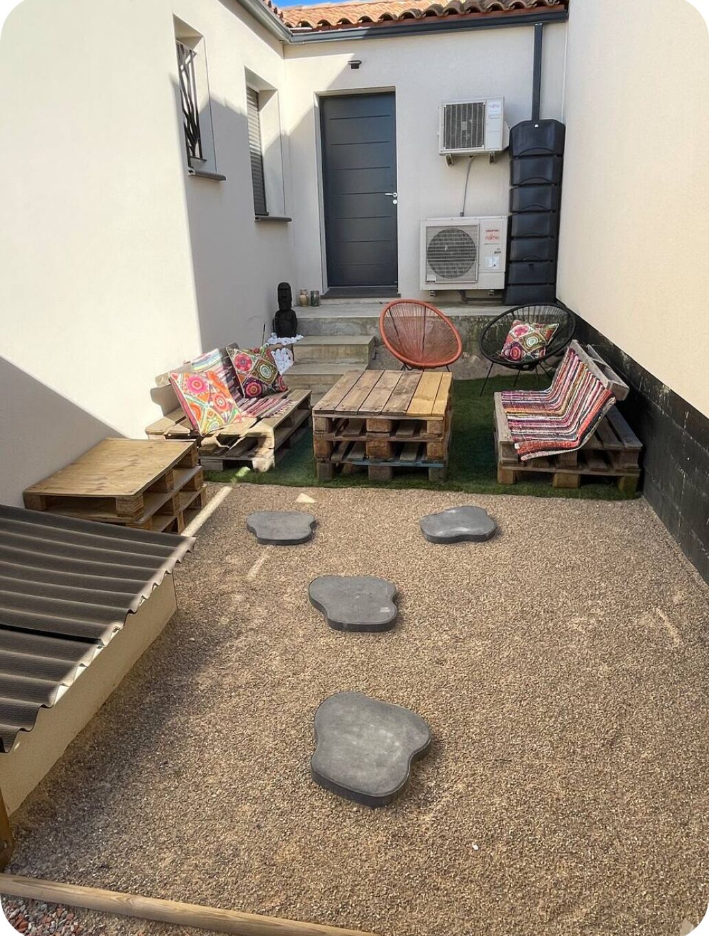 Un patio con muebles de madera, una mesita y una silla. Hay escalones que conducen a una puerta, y se ve un equipo de aire acondicionado en la pared.