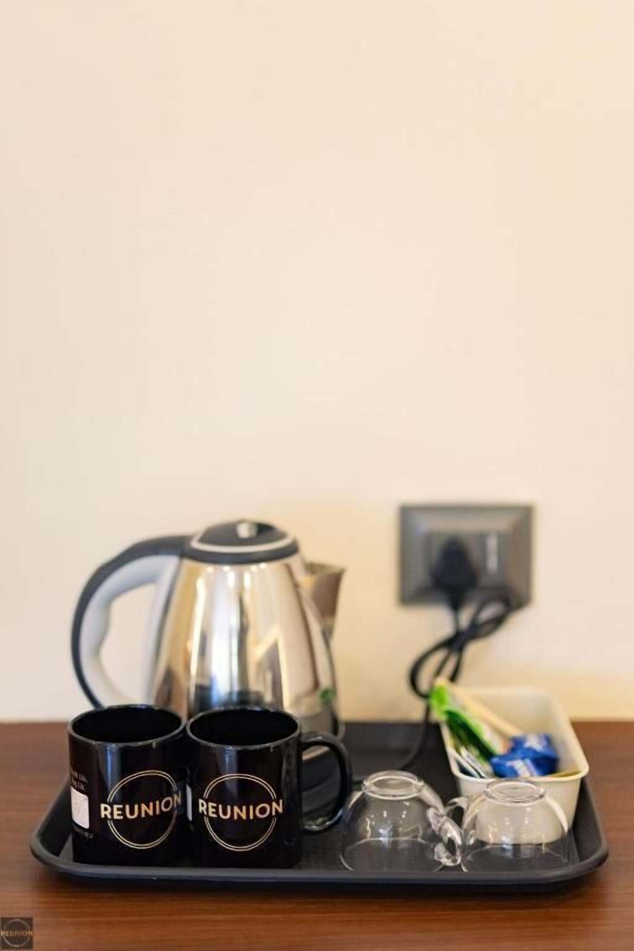 A tray with two black mugs labeled 'REUNION,' a silver teapot, and a small glass container with a green object on a wooden table against a plain wall.