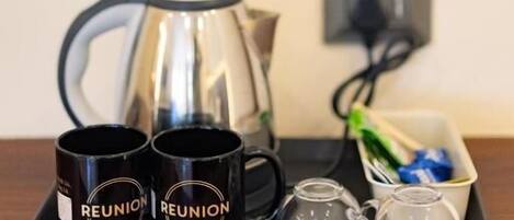 A tray with two black mugs labeled 'REUNION,' a silver teapot, and a small glass container with a green object on a wooden table against a plain wall.