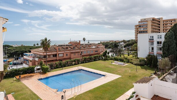 Piscina cercada por uma área gramada, um edifício com telhado vermelho e vista para o mar.