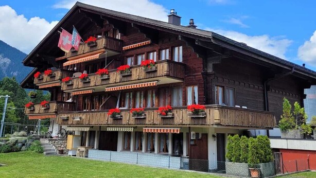 A multi-story wooden building with balconies, a Swiss flag, and a mountain backdrop. - Apartment Anouk Centrally located, cozy family apartment with sunny terrace (Lenk)