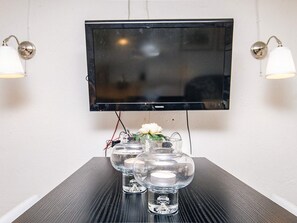 A flat-screen TV mounted on a wall, two wall-mounted lamps, and a table with three clear glass vases with flowers.