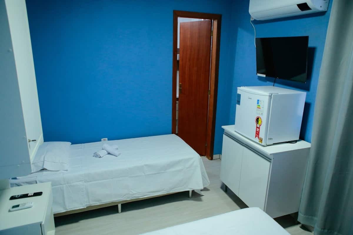 Una habitación pequeña y limpia con una cama individual, un escritorio, un refrigerador y un televisor fijado a la pared.