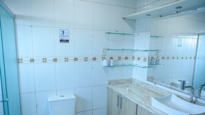 Un baño con inodoro, lavamanos y espejo. Hay repisas de vidrio sobre el lavamanos y un letrero en la pared.