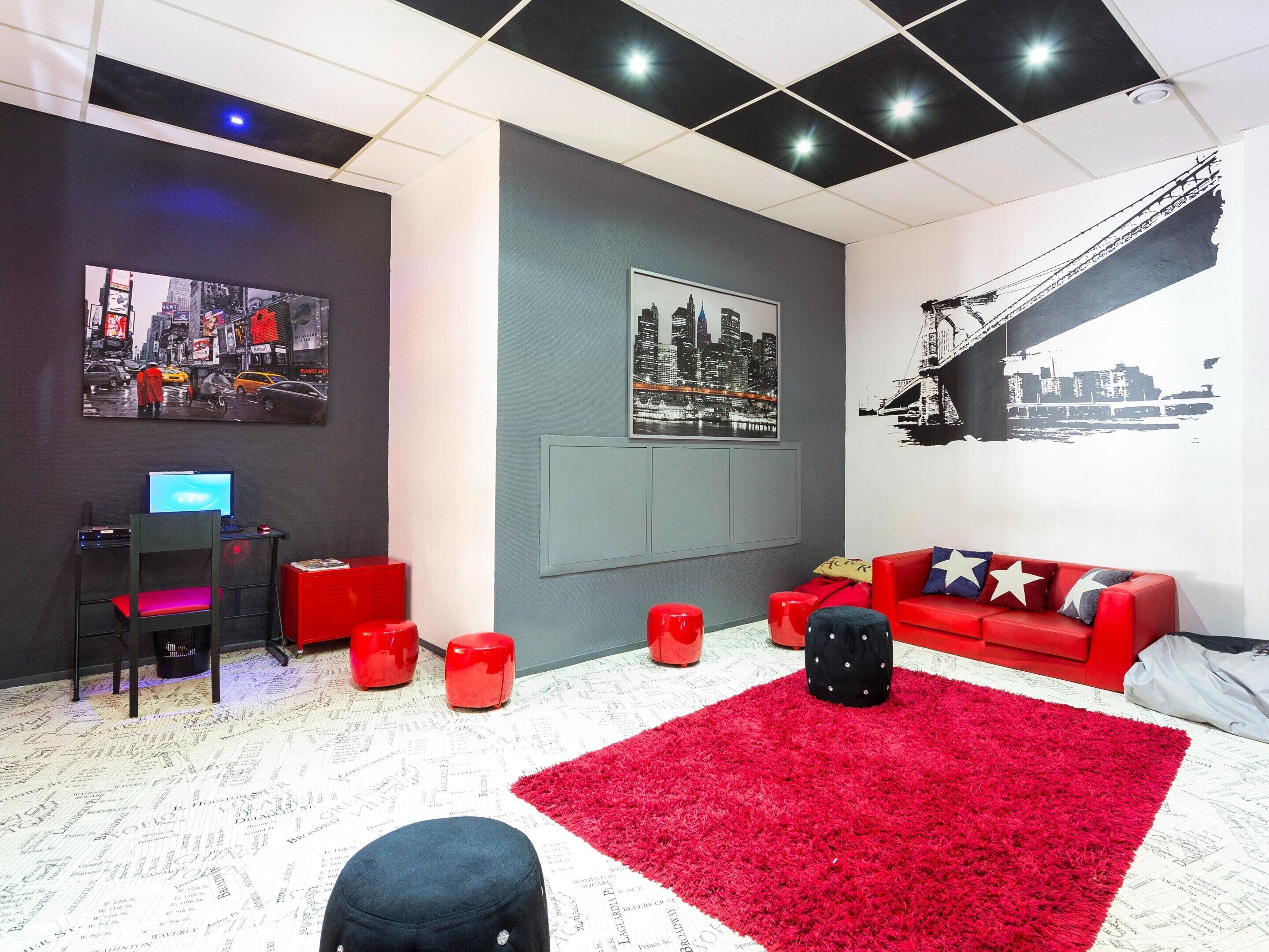 A modern living room with a red sofa, a red ottoman, a black stool, a red desk with a laptop, and a cityscape mural on the wall.