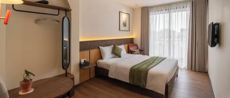 A hotel room with a bed, bedside table, lamp, potted plant, and a large window with curtains.