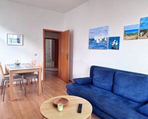 A living room with a blue sofa, a dining table with chairs, and artwork on the walls. - Berta`s Heim Meerblickappartement (Sassnitz)