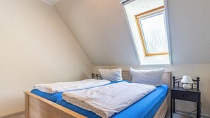 Quarto com teto inclinado, um claraboia, cama com roupa de cama azul, almofadas brancas e mesa de cabeceira com abajur.